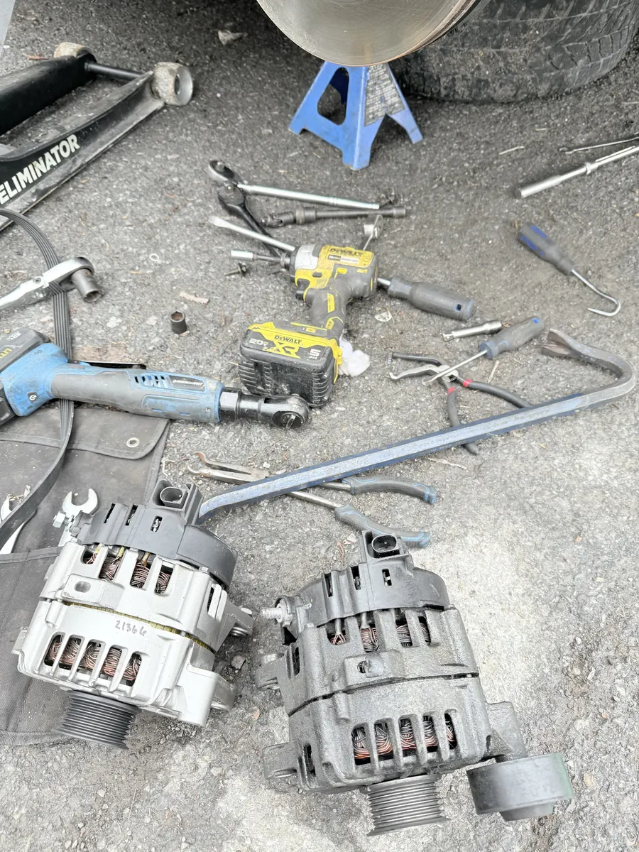 Old and new alternators on the ground next to tools and a jack stand