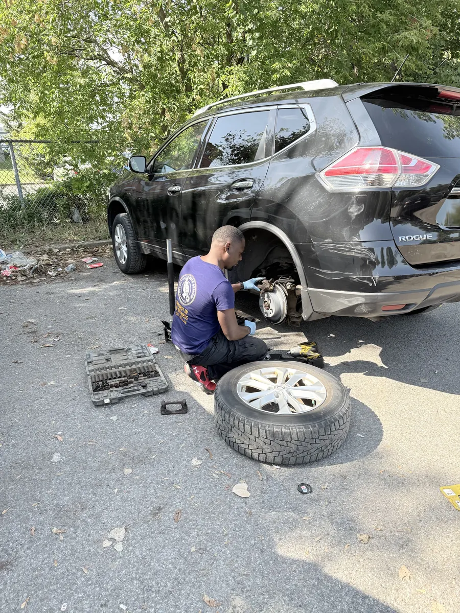 Elie in an Alimotorsforall shirt kneeling at the rear brake of a black Nissan Rogue, wheel off, tools beside him